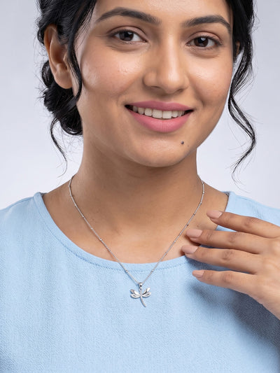A close-up of a woman wearing a delicate silver necklace with a small dragonfly pendant, paired with a light blue top against a neutral gray background.